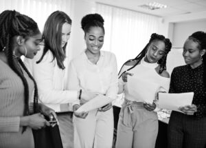 Five multiracial business womans standing at office with papers Five multiracial business womans standing at office with papers on hands. Diverse group of female employees in formal wear.
